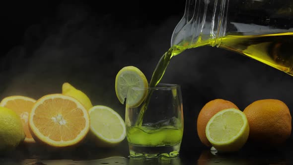 Pour Juice From Pitcher Into Glass with Ice, Oranges and Lemons Background
