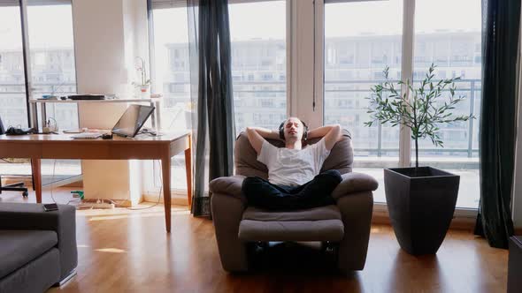 Young man sitting in armchair listening music with headphones