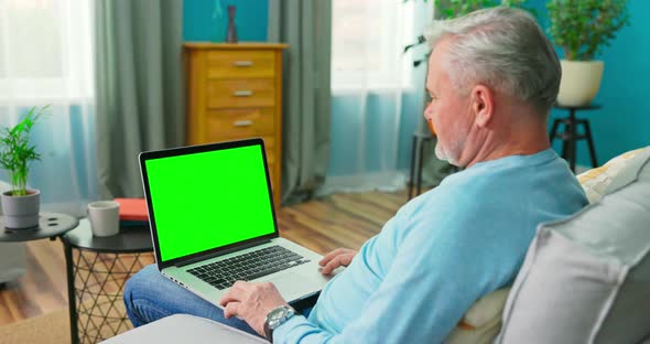 Older Man at Home Sitting on a Couch Works on a Laptop Computer with Green