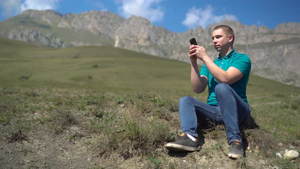 A Young Man Sits with a Phone in His Hands in the Mountains. The Man Is in Correspondence of the