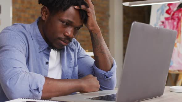 Stressed african american male artist looking at his laptop while sitting on his desk at art studio