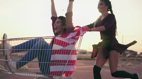 Young Hipster Teen Girls Having Fun at the Shopping Mall Parking Riding in Shopping Cart Holding the