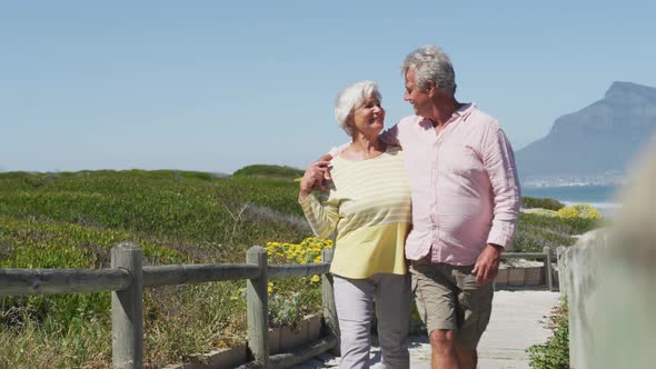 Happy senior caucasian couple with arms around each other walking on path leading to the beach