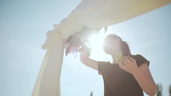 Young Female with Brown Hair Decorating Wedding Arch with Flowers Outdoors While Sun Shines Right at