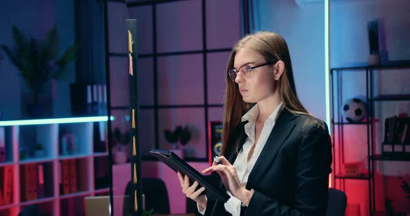 businesswoman in glasses standing near glass wall and writing out important datas from tablet pc