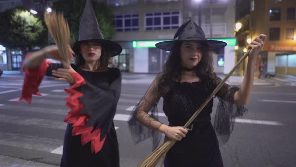 Two Colombian Girls In Witch Halloween Costumes With Broom Sticks Looking At Camera Smiling While