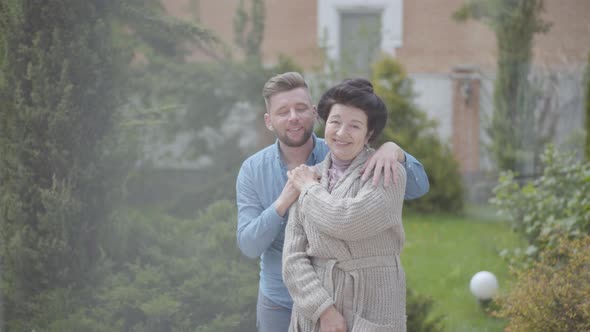 Portrait Happy Mature Woman Standing in the Garden in Front of the Big House, Adult Grandson Hugging