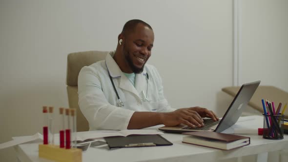 Cheerful Black Male Physician in Hands Free Earphones Enjoying Music While Working on Laptop