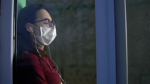 A Young Woman Is Sitting at Home in Quarantine with Headphones in Her Ears. A Girl Sits on a Window