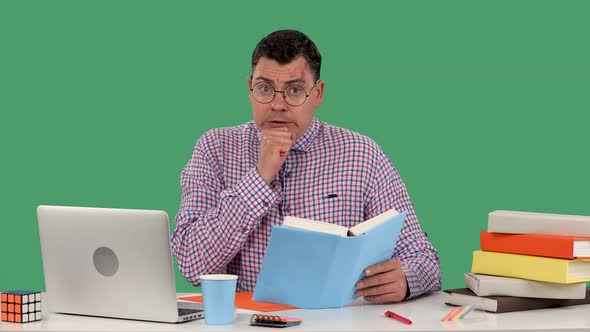 A Man with Glasses Sits at a Desk in Front of a Laptop and Reads a Book Shudders Puts His Fist to