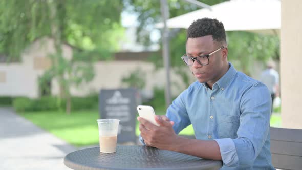 African Man Celebrating Success on Smartphone in Outdoor Cafe