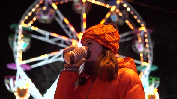 Girl Drinks Tea on the Background of the Ferris Wheel