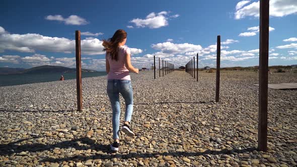 Young and Attractive Girl or Young Woman in Pink T-shirt Runing on the Beach Between Wood Pillars