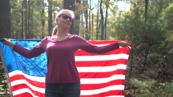 Smiling, blonde woman walking through the forest wrapping an American flag around her.