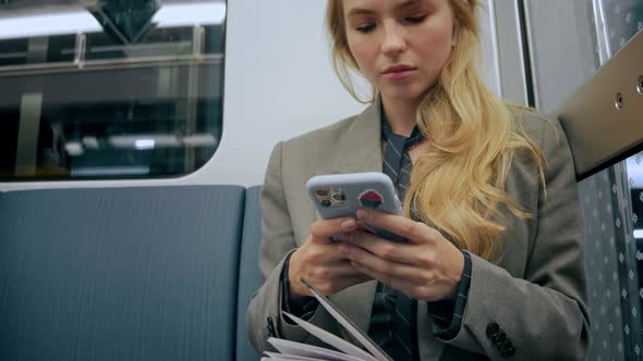 Young Blonde Business Girl Sitting in Modern Metro and Looking at Her Phone