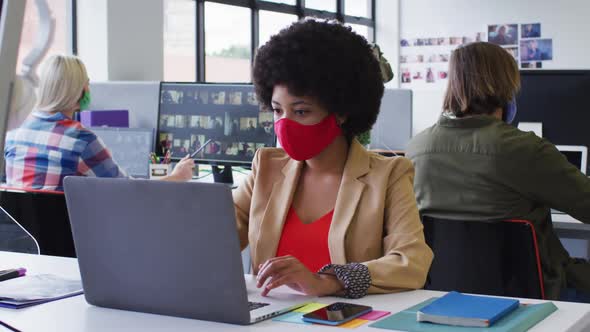 Mixed race businesswoman wearing face mask sitting using a laptop in modern office