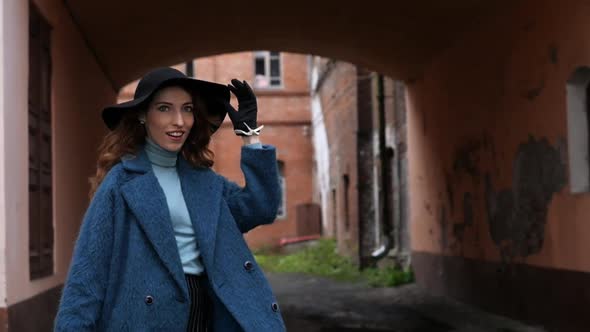 Young Redhead Woman in a Blue Coat and Black Hat with a Suitcase on the Background of the Old City