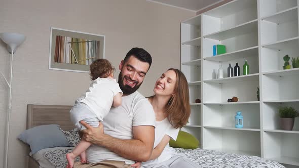 Young Bearded Father Raises Daughter Above His Head, Mother Sitting on Bed and Looks at Family