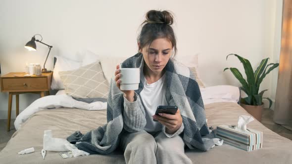 Sick Young Woman with Hot Drink Using Smartphone in Bed