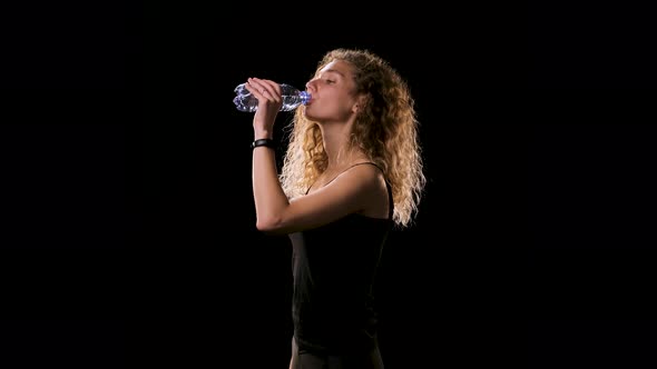 Beautiful Blonde Hair Lady with Lush Wavy Hair on Black Background in Black Top Drinks Water From