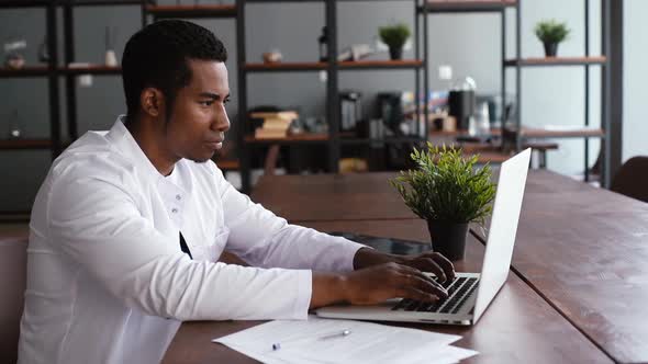 Focused Young African Male Doctor in White Coat Working Typing on Laptop Computer Sitting at Desk.