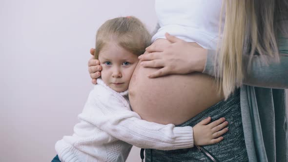 Pregnant Woman with Belly Hugs Little Daughter at Light Wall