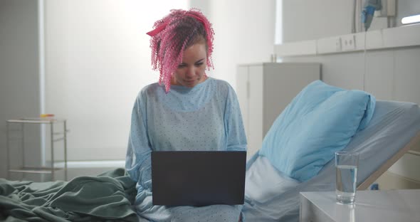 Young Female Patient Working with Laptop Sitting on Bed in Hospital Room