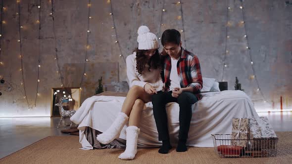 Happy Young Couple Checking Christmas Messages on a Smartphone Sitting on the Bed at Home