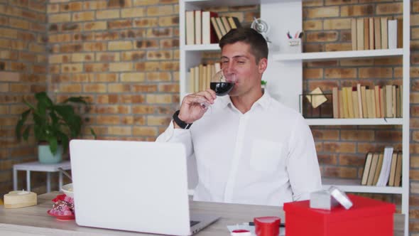 Caucasian man making video call using laptop drinking glass of wine and talking