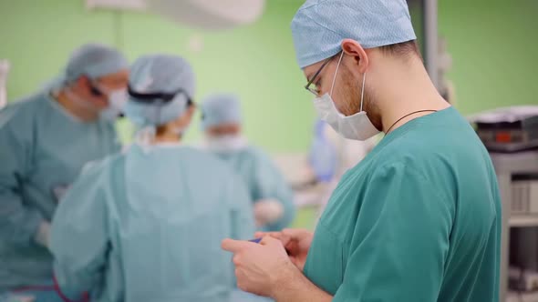 Assistant in Surgery Is Swiping Pages on Screen of Smartphone, Team of Surgeons Are Working