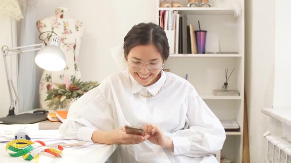 Happy Designer Girl with a Smartphone Sitting in Home Workshop