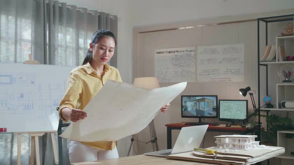 Asian Woman Engineer With A Laptop Looking At Blueprint While Working At The Office