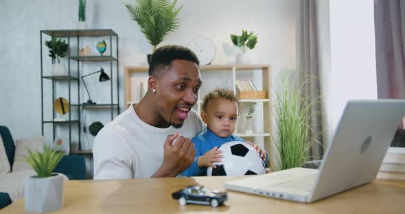 Dad Holding His Calm Baby Boy on Knees During watch Football Game on Computer