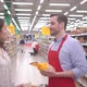 Male stock clerk in red apron, helping young attractive woman to choose fruits in grosery store - VideoHive Item for Sale