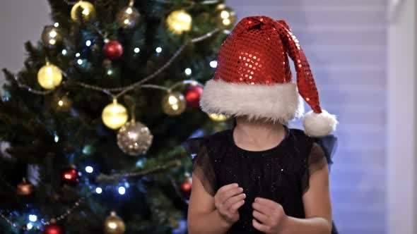 Child girl in Santa hat play hide and seek near a Christmas tree. Playful mood