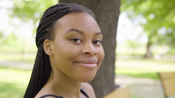 A Young Black Woman Turns and Smiles at the Camera As She Sits on a Bench in a Park on a Sunny Day