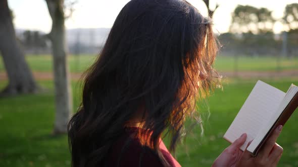 A young woman reading a story book outdoors in the park at sunset with light shining bright.