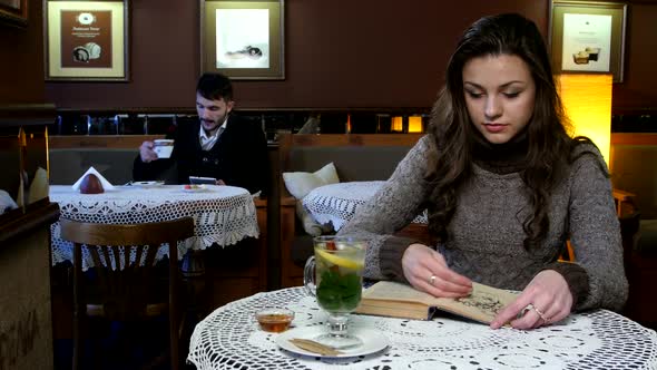 Boy and Girl Sitting in Beautiful Cafe. He`s Reading Something Into His Tablet Computer, She Reads a
