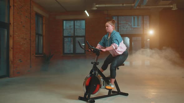 Athletic Woman Riding on Spinning Stationary Bike Training Routine in Haze Gym Weight Loss Indoors