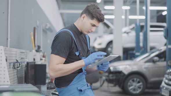 Side View Portrait of Concentrated Young Auto Mechanic Surfing Internet on Tablet