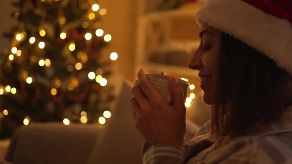 Woman in Pajama and Red Santa Hat Sitting on Couch Watching TV and Drinking Hot Chocolate From Mug