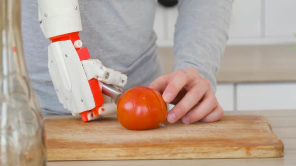 Man with Robotic Prosthetics Hand with Knife on It Is Cutting Tomato on Wooden Board