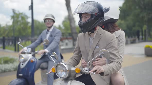 Portrait of Confident Caucasian Man in Formal Suit and Helmet Sitting on Retro Scooter with Girl on