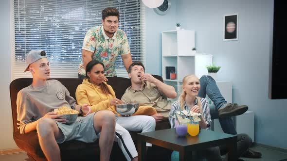 Excited Friends Relaxing on Sofa Watching TV and Eating Snacks at Home