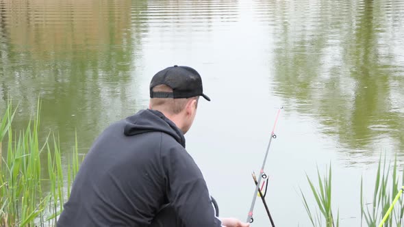 A Man Fishes Stands on the Shore of a Large and Calm Lake and Catches Fish with a Fishing Rod and