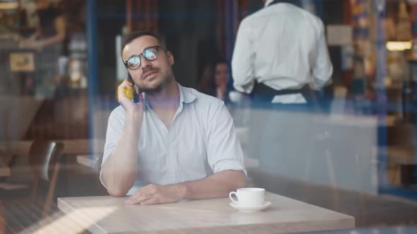 Smiling Handsome Man Entrepreneur Talking To Client on Smartphone at Cafe Drinking Coffee