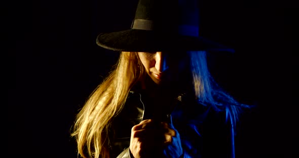 Portrait of a Blonde in a Hat on a Black Background. She Looks Down, Pulls Her Jacket Tighter Around