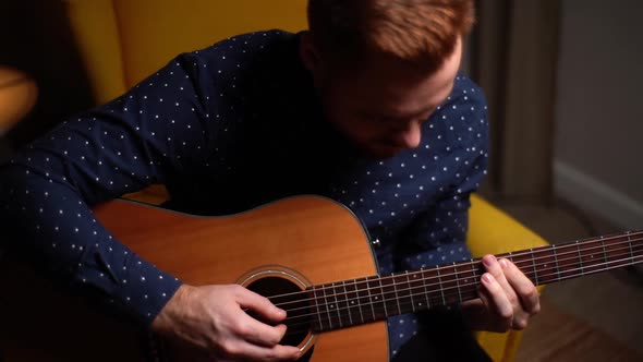 Closeup Top View of Talented Young Guitarist Man Playing Guitar Sitting on Armchair in Dark Living