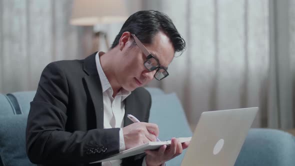 Asian Businessman In Jacket And Shorts Writing In A Notebook While Working With A Laptop At Home