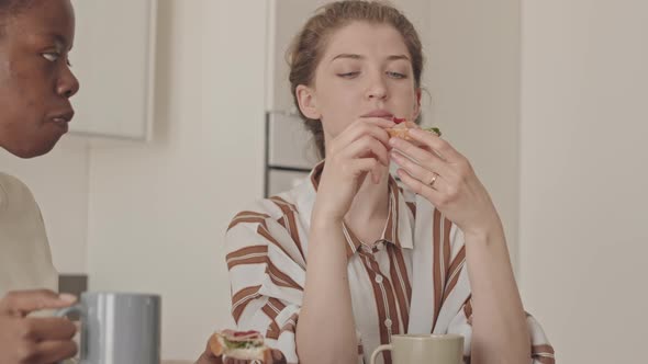 Two Diverse Girlfriends Eating Homemade Sandwiches at Kitchen Table